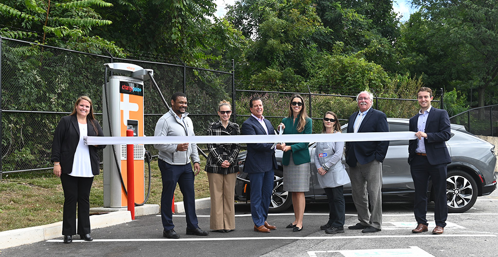 Eight people standing behind a long white ribbon in a parking lot. An electric vehicle charging station and car are behind them.