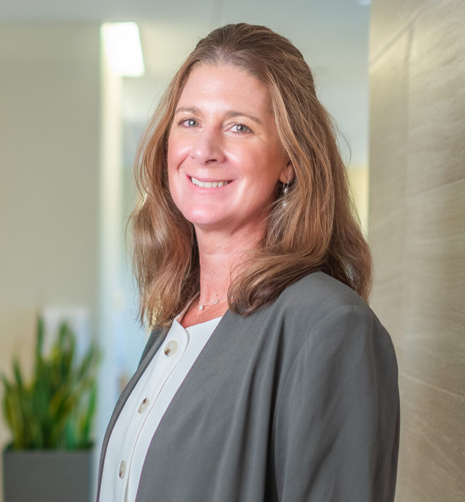 Person wearing a gray blazer over a white buttoned top, standing indoors near a light-colored wall with a green plant in the background.