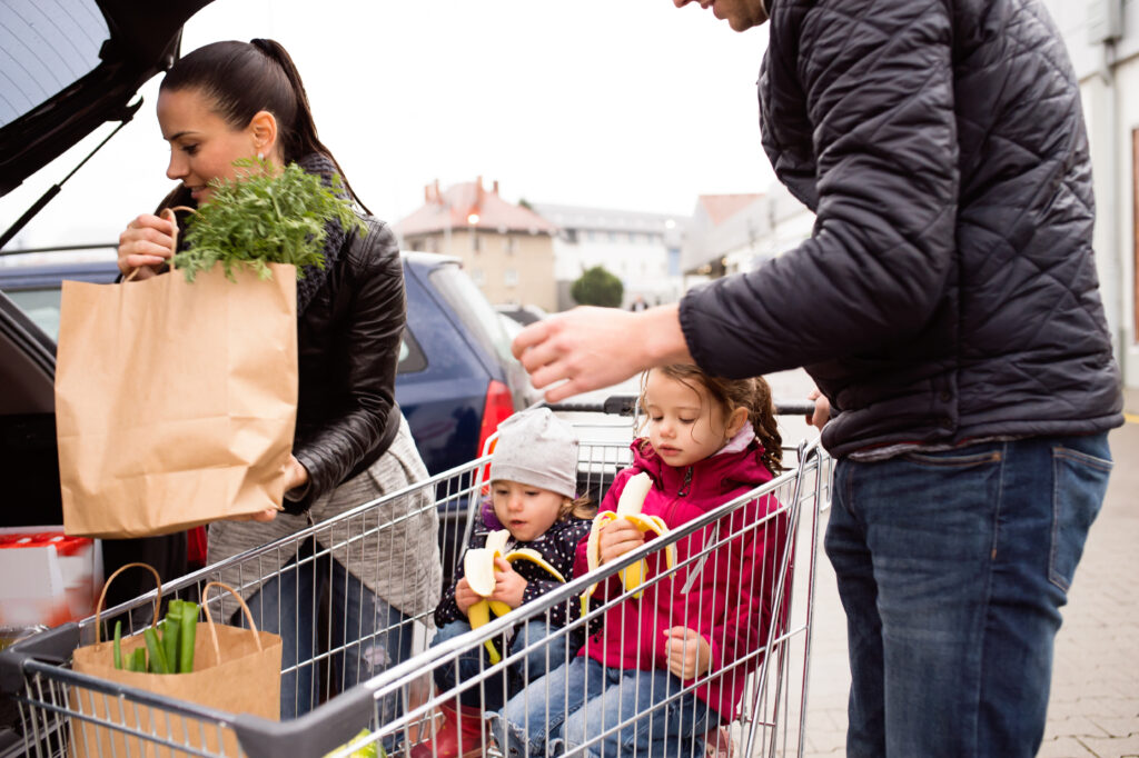 Family filling car with fresh healthy groceries, two children sitting in grocery cart enjoying eating fresh bananas.