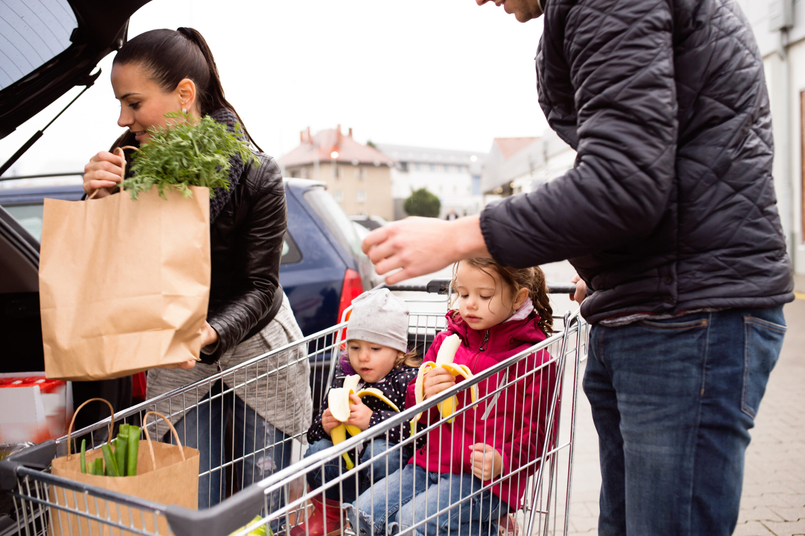 Family filling car with fresh healthy groceries, two children sitting in grocery cart enjoying eating fresh bananas.