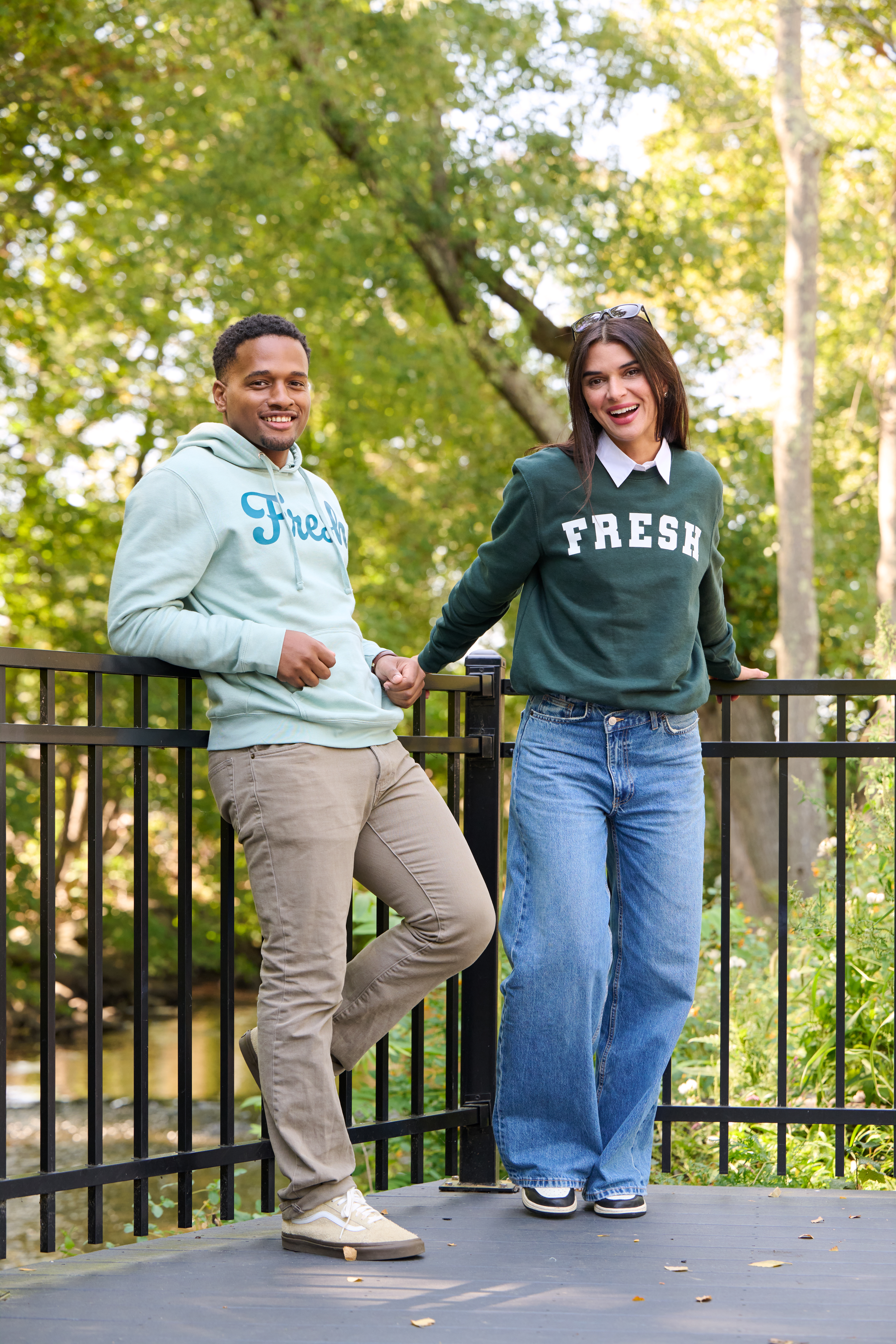 Two people standing outdoors on a black metal railing platform surrounded by trees, wearing casual outfits with green ‘Fresh’ sweatshirts and jeans.
