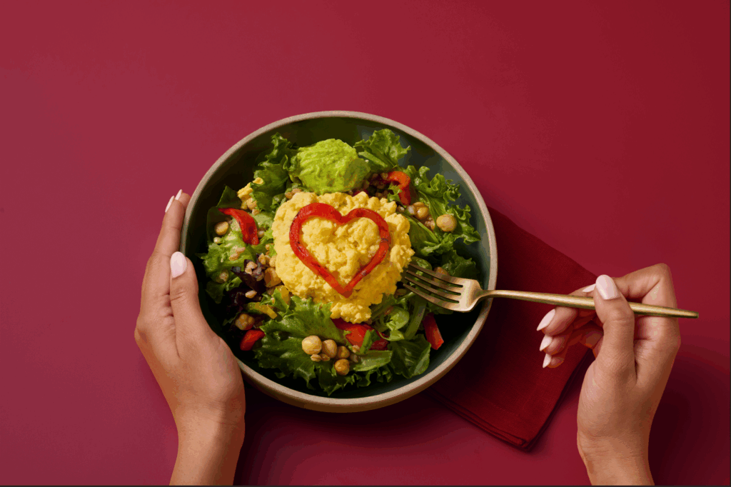 Bowl of salad topped with a mound of scrambled eggs shaped with a red pepper heart, with hands holding a fork.