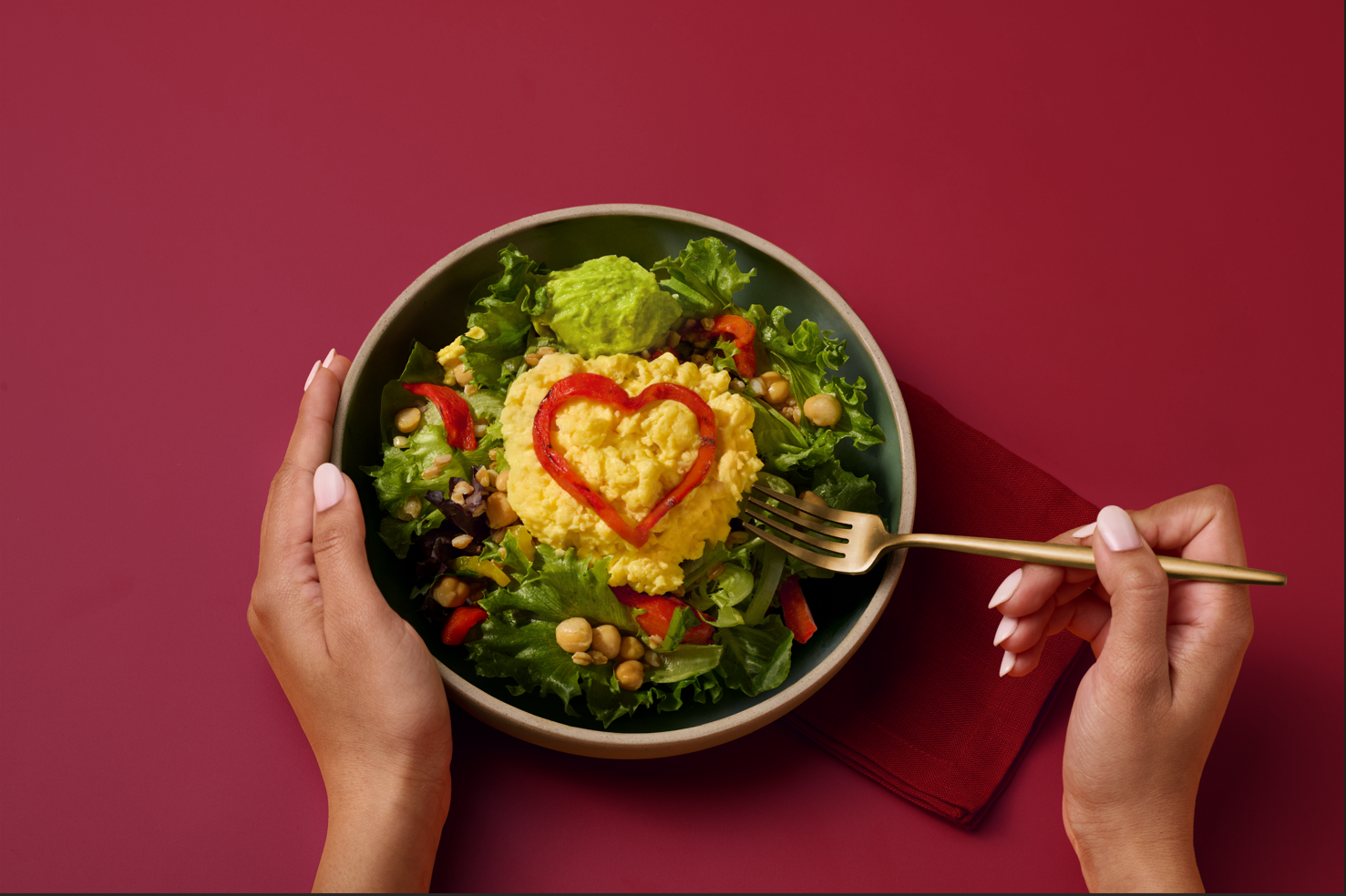 Bowl of salad topped with a mound of scrambled eggs shaped with a red pepper heart, with hands holding a fork.