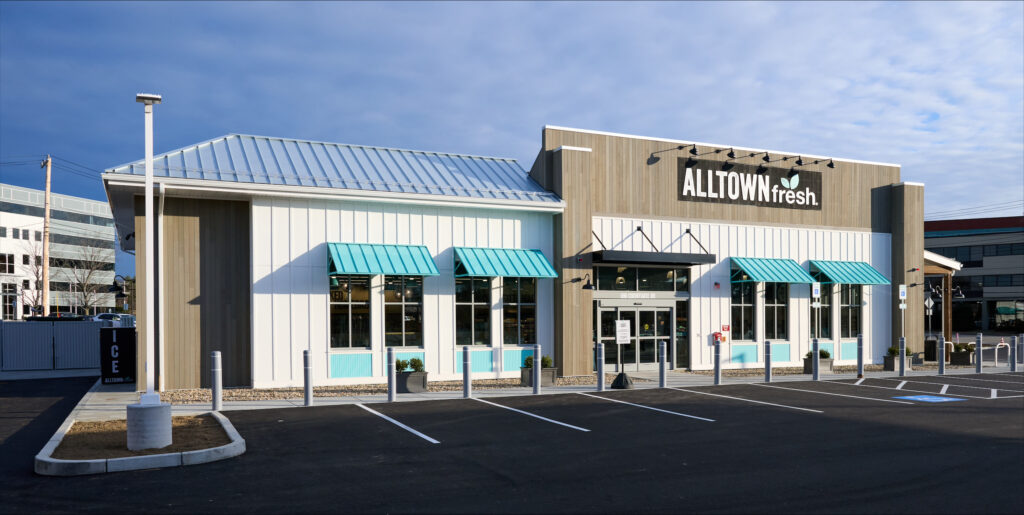 Exterior of a convenience store with a light exterior, teal window awnings, and a large “ALLTOWN fresh” sign above the entrance, shown with an empty parking lot in front.