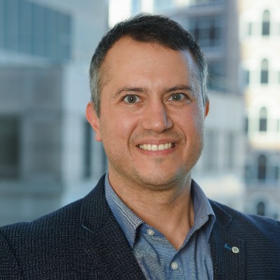 Person wearing a dark blazer over a blue collared shirt, photographed indoors with a softly blurred city background.