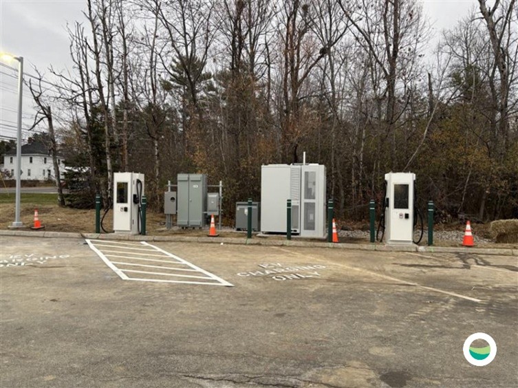 Outdoor electric vehicle charging station with multiple chargers and equipment installed along a paved lot, bordered by trees in the background.