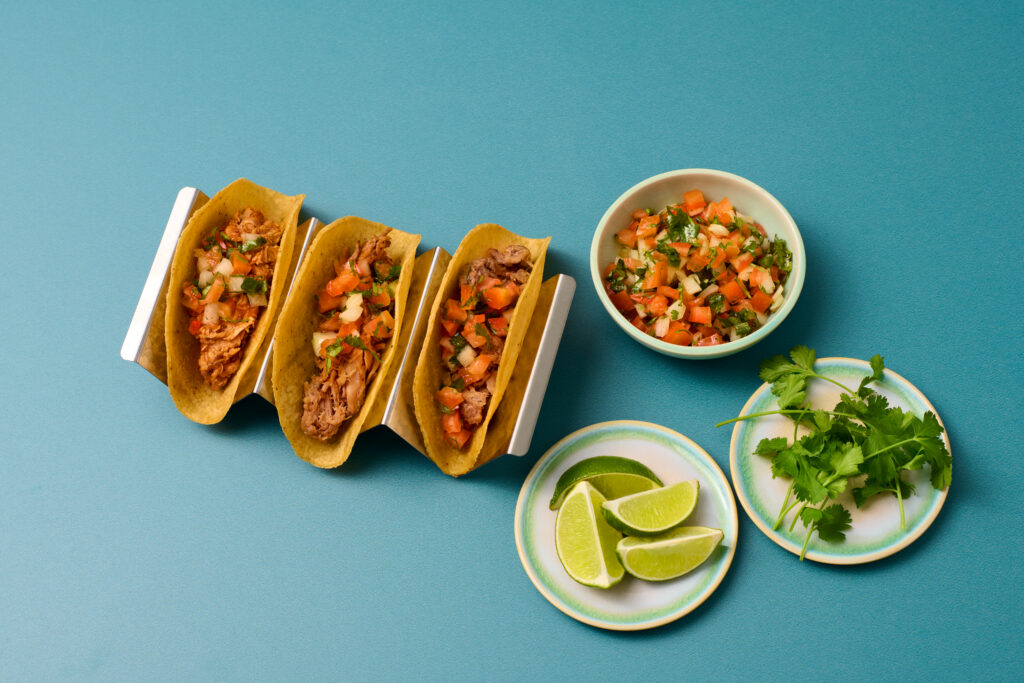 Three tacos on a blue surface with small bowls of salsa, guacamole, and lime wedges.