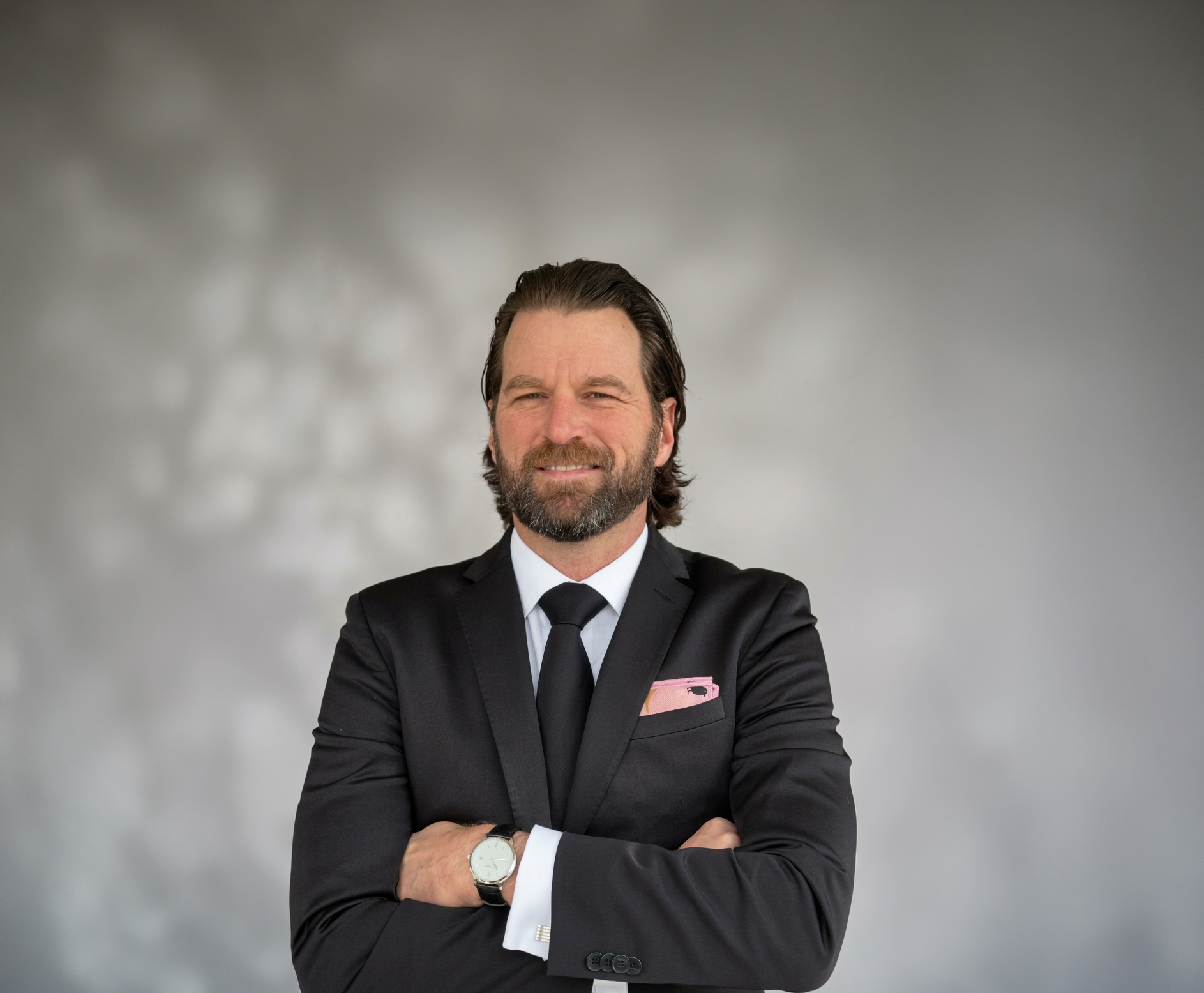 Person wearing dark suit with pocket square, crossing arms.